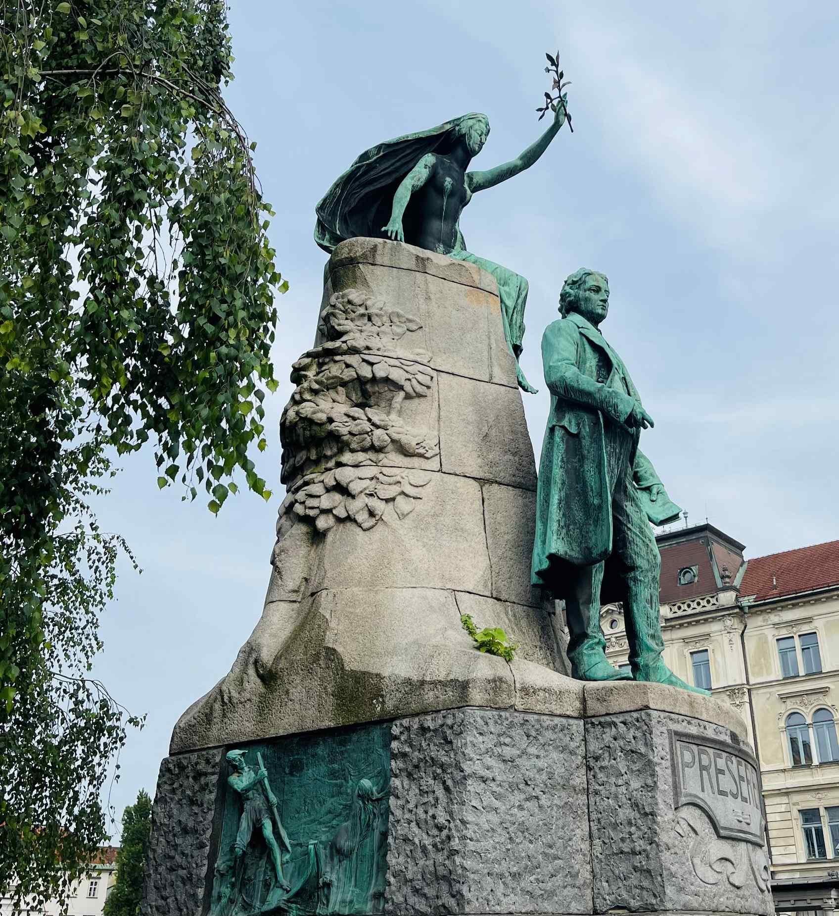 Statues of angelic figure holding a branch atop a stone structure with man in dress coat standing in front in Ljubljana
